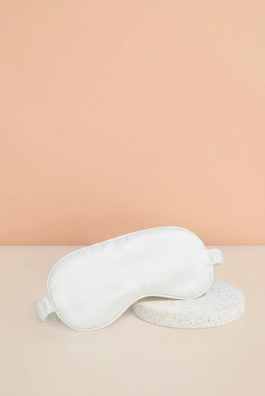 A white sleep mask with the word 'Bride' embroidered in pink script, resting on a round textured white coaster against a peach and beige background.